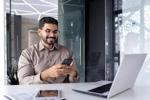 homem de negocios segurando telefone dentro escritório, alegre homem sorridente usa Smartphone aplicativo às ambiente de trabalho, navega social redes, e escreve texto mensagem. foto