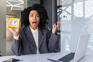 sobrecarregado mulher trabalhando tarde, empresária olhando às Câmera chateado mostrando relógio, fêmea trabalhador com computador portátil dentro meio do escritório cansado. foto