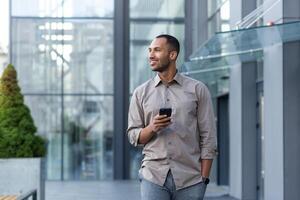 sorridente hispânico homem caminhando baixa rua perto moderno escritório prédio, trabalhador autonomo homem de negocios olhando longe segurando Móvel telefone foto