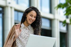 uma confiante empresária com uma computador portátil é acenando Olá lado de fora uma moderno escritório prédio, incorporando profissional elegância e simpatia. foto