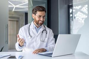 uma sorridente jovem masculino médico senta dentro a escritório do a clínica às uma mesa, comunica e consulta conectados através uma computador portátil. foto