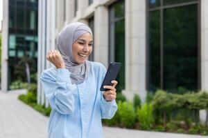 alegre mulher dentro hijab caminhando dentro a cidade, recebido notificação do conectados ganhar e Boa notícia em telefone, o negócio mulher lado de fora escritório prédio, segurando mão acima triunfo vencedora gesto. foto