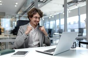 uma jovem empreendedor com encaracolado cabelo mostra excitação dentro a escritório, manifestando sucesso às dele local de trabalho com uma computador portátil dentro frente. foto