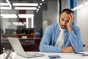 chateado e cansado jovem hispânico homem trabalhando dentro uma confuso escritório sentado taciturnamente às uma escrivaninha com uma computador portátil, pensativamente em repouso dele cabeça em dele mão. foto