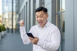 agradavelmente chocado jovem masculino ásia homem de negocios em pé perto a escritório, olhando alegremente às a Móvel telefone tela e alegria dentro sucesso mostrando uma vitória gesto com dele mão. foto