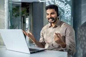 jovem bem sucedido homem de negocios às local de trabalho falando remotamente sentado às mesa dentro escritório, homem com fone de ouvido usando computador portátil para chamar, conectados cliente Apoio, suporte trabalhador. foto