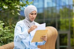 uma preocupado muçulmano mulher vestindo uma hijab segurando e lendo uma documento ou carta enquanto sentado lado de fora em uma banco. foto