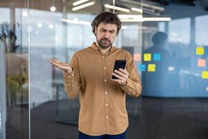 uma meia idade homem de negocios parece confuso e insatisfeito Como ele usa dele Smartphone dentro uma moderno escritório ambiente, cercado de pegajoso notas em vidro paredes. foto