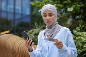 chateado enganado mulher com telefone tentando para faço conectados compra e dinheiro transferir, muçulmano mulher dentro hijab desapontado obteve rejeição, segurando telefone e banco crédito cartão, insatisfeito comprador. foto