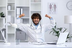 bem feito Super heroi doutor, homem sorrisos parece às Câmera, detém dele mãos acima, regozija-se às a sucesso dentro a tratamento do a paciente, trabalho dentro a clínica dentro a médico escritório, senta às a mesa. foto