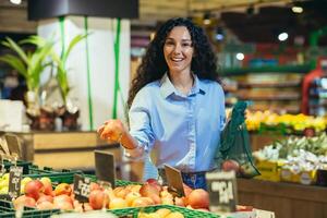 retrato do feliz mulher comprador dentro supermercado, hispânico mulher escolhe maçãs e frutas sorridente e olhando às Câmera, com mercearia cesta escolhe bens foto