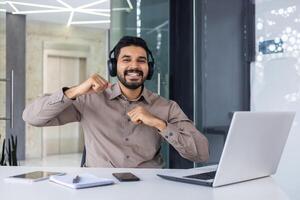 retrato do uma feliz jovem indiano homem sentado às uma escrivaninha dentro a escritório, trabalhando em uma computador portátil, olhando sorridente às a Câmera e dançando dentro alegria do sucesso. foto