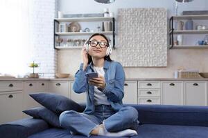 jovem lindo ásia mulher relaxante às casa sentado em sofá dentro vivo sala, fêmea aluna ouvindo para música dentro fones de ouvido e usando aplicativo em telefone para ouço para conectados rádio e podcasts foto