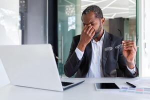 sobrecarregado homem às local de trabalho dentro escritório, africano americano homem de negocios trabalhando sentado com computador portátil às trabalhar, patrão levando fora óculos e massageando olhos. foto