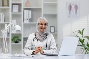 uma alegre fêmea médico vestindo uma hijab dentro uma moderno clínica escritório, sentado às dela mesa, usando uma computador portátil, e olhando às a Câmera com uma amigáveis sorriso. foto