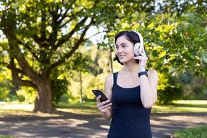 jovem sorridente desportivo mulher indo para manhã corre e exercício. em pé dentro a parque vestindo fones de ouvido e segurando uma telefone. foto