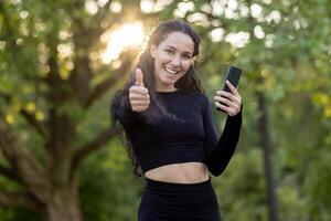 alegre Atlético mulher com Smartphone dentro natureza mostrando aprovação com polegares acima durante dourado hora. foto