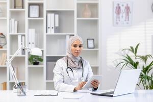 profissional muçulmano fêmea médico dentro uma hijab e branco casaco trabalhando às dela escritório, usando uma tábua, com computador portátil e estetoscópio em escrivaninha. foto