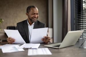 africano americano homem de negocios dentro uma terno sorrisos enquanto revendo papéis dentro a escritório configuração com uma computador portátil e de outros documentos em dele escrivaninha. foto