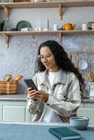 vertical tomada, lindo hispânico mulher usando Smartphone às lar, mulher com encaracolado cabelo e óculos sorridente, digitando mensagem e lendo notícia on-line, aluna dentro cozinha. foto