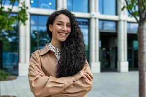 jovem lindo hispânico mulher com encaracolado cabelo lado de fora escritório prédio, companhia trabalhador sorridente e olhando às Câmera com cruzado braços, satisfeito empresária. foto