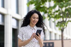 uma jovem lindo mulher anda em através a cidade com uma telefone dentro dela mãos, sorrisos contente, usa uma Smartphone aplicativo, navega Internet Páginas, tipos uma texto mensagem, social redes. foto