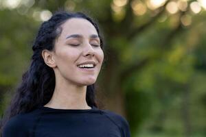 uma hispânico mulher fecha dela olhos com uma conteúdo sorriso, aquecendo dentro serenidade no meio uma natural verde pano de fundo. foto