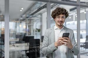 uma jovem homem de negocios com encaracolado cabelo Verificações dele Smartphone dentro uma bem iluminado, contemporâneo escritório ambiente, exalando profissionalismo e foco. foto