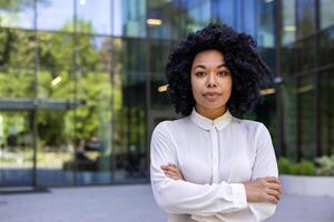 sério Preto mulher dentro branco camisa em pé com cruzado braços em peito lado de fora do vítreo construção com refletindo árvores corporativo meio era Empregador olhando às Câmera com orgulhoso expressão. foto