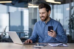 focado profissional trabalhando em uma computador portátil e segurando uma Smartphone dentro uma moderno escritório contexto, expressando produtividade e compromisso. foto