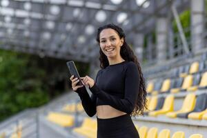 uma sorridente jovem mulher vestindo Atlético vestem detém uma Smartphone enquanto em pé às uma Esportes estádio com assentos dentro a fundo. foto