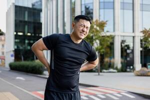 jovem ásia masculino atleta segurando dele costas e sentindo-me forte dor. em pé em uma cidade rua e sofrimento a partir de dor. foto