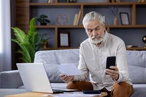 focado idosos homem com branco cabelo e barba, revendo papéis e verificação em formação em dele Smartphone enquanto sentado em uma sofá. uma confortável casa configuração com computador portátil e interior plantas. foto