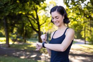 feliz jovem mulher é fazendo Esportes e corrida dentro a parque, em pé olhando às a inteligente Assistir e feliz com a resultado mostrando uma sim gesto com dela mão. foto