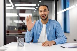 alegre indiano homem de negocios dentro uma casual azul camisa noivando dentro uma ligar a partir de dele moderno escritório, cumprimento com uma aceno. foto