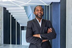 retrato do bem sucedido africano americano chefe, homem de negocios dentro o negócio terno sorridente e olhando às Câmera com cruzado braços dentro escritório, homem satisfeito com trabalhos e conquistas. foto