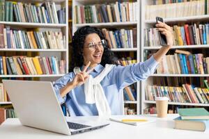 alegre hispânico fêmea aluna leva uma brincalhão selfie entre livros dentro uma biblioteca contexto, exemplificando aluna vida e felicidade. foto