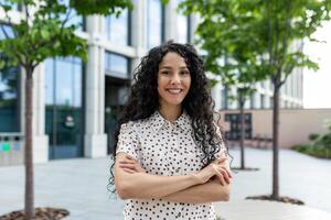 jovem lindo hispânico mulher com encaracolado cabelo sorridente e olhando às Câmera, empresária com braços cruzado lado de fora escritório construção ao ar livre. foto