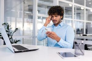 problemas com a conta, aplicativo, conectados compras, mau mensagem. uma jovem latim americano homem é sentado às uma escrivaninha dentro a escritório, segurando dele cabeça preocupado, olhando Infelizmente às a telefone ele é usando. foto