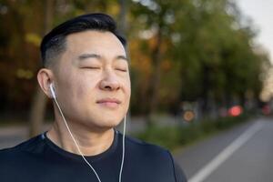 fechar-se foto do uma jovem ásia homem dentro fones de ouvido em pé em uma cidade rua, fazendo Esportes, fechadas dele olhos, em repouso, desfrutando música.