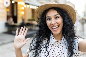 jovem lindo hispânico mulher sorridente e olhando para dentro telefone Câmera, viajante usa Smartphone aplicativo para controlo remoto comunicação chamar, anda em dentro tarde cidade, em turista Tour viagem. foto