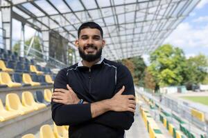 uma jovem homem sorrisos com confiança com braços cruzado, em pé dentro a esvaziar ao ar livre estádio com amarelo assentos e uma Claro céu. foto