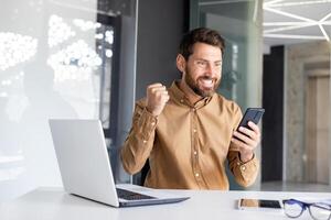 homem vencedora segurando telefone dentro mãos, homem de negocios às local de trabalho feliz com conectados realização resultados, trabalhador usando aplicativo em Smartphone, sentado dentro escritório com computador portátil, vestindo casual camisa. foto