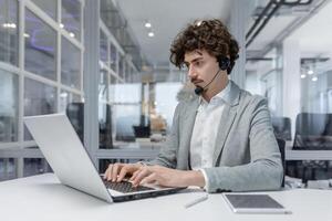 uma sério jovem homem de negocios homem dentro uma fone de ouvido é sentado dentro a escritório às uma escrivaninha e é concentrando em trabalhando em uma computador portátil. foto