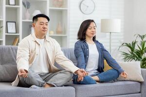 feliz ásia família às casa em sofá meditando, casal homem e mulher sentado dentro lótus posição dentro vivo quarto com fechadas olhos em repouso relaxante junto. foto