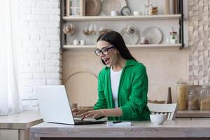 uma jovem mulher é sentado às casa dentro a cozinha usando uma computador portátil. tocam conectados jogos, loteria, sorteio foto