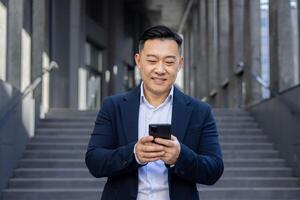 sorridente ásia executivo usando Móvel telefone para mensagens de texto enquanto em pé em escadas lado de fora uma moderno prédio. foto