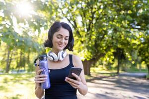 focado em fitness fêmea goza uma pausa dentro a parque, verificação dela telefone com água garrafa e fones de ouvido. foto