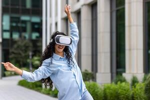 jovem lindo menina dentro camisa e jeans dançando lado de fora escritório construção vestindo virtual realidade copos, hispânico mulher feliz tendo Diversão e relaxante com encaracolado cabelo. foto