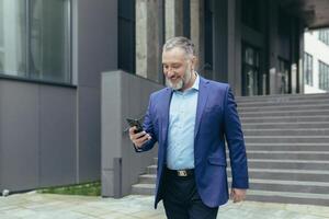 Senior grisalho bonito homem dentro uma terno. ele anda em perto a escritório prédio, detém a telefone dentro dele mãos, faz uma chamar, fala, sorri. foto